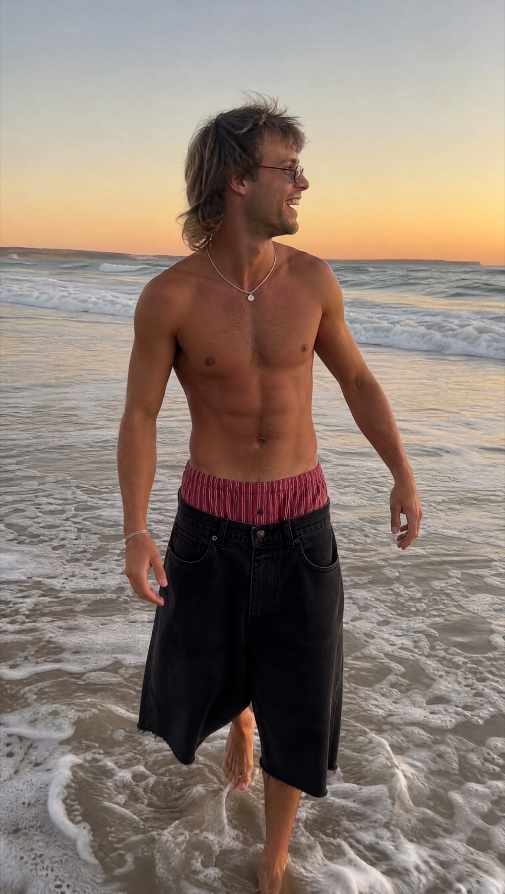 Model at the beach with a red striped boxer waistband visible above black denim shorts